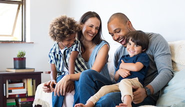 Happy multiethnic family sitting on sofa laughing together. Cheerful parents playing with their sons at home. Black father tickles his little boy while the mother and the brother smile.