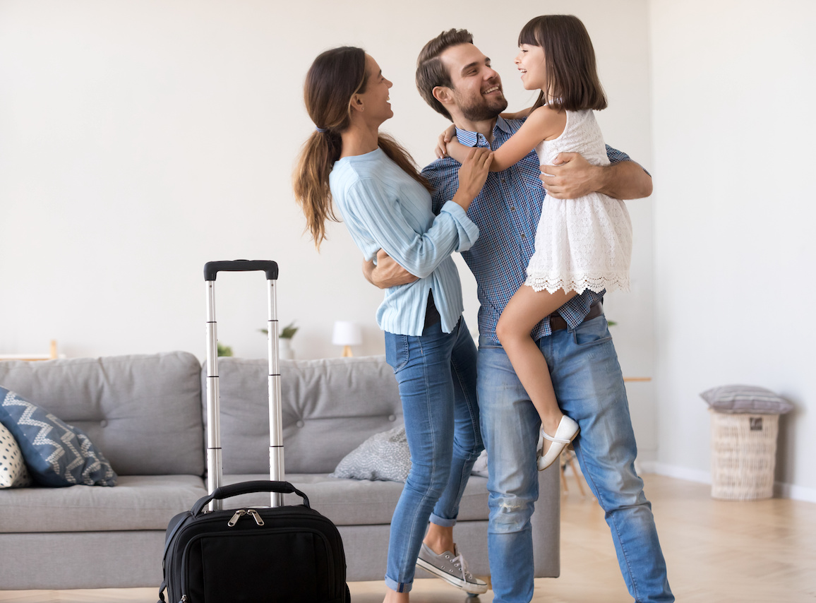 Happy father embracing child and young wife returning home with suitcase.